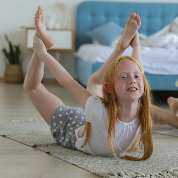 Full body of cheerful girl in casual clothing with reddish hair looking away with smile while stretching legs on carpet in bright cozy living room on blurred background